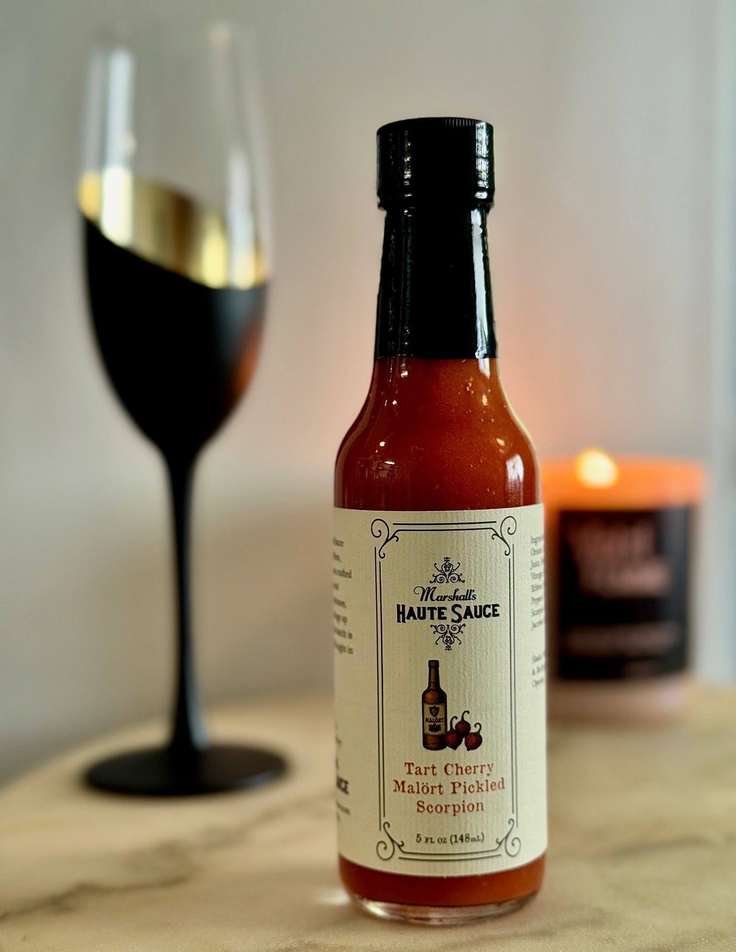 Tart Cherry Malört Hot Sauce bottle next to a wine glass and candle, showcasing bold flavor and spice.