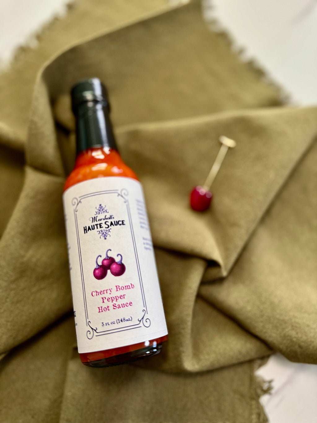 Cherry Bomb Pepper Hot Sauce bottle on olive green fabric, showcasing bold flavors and vibrant cherry design.