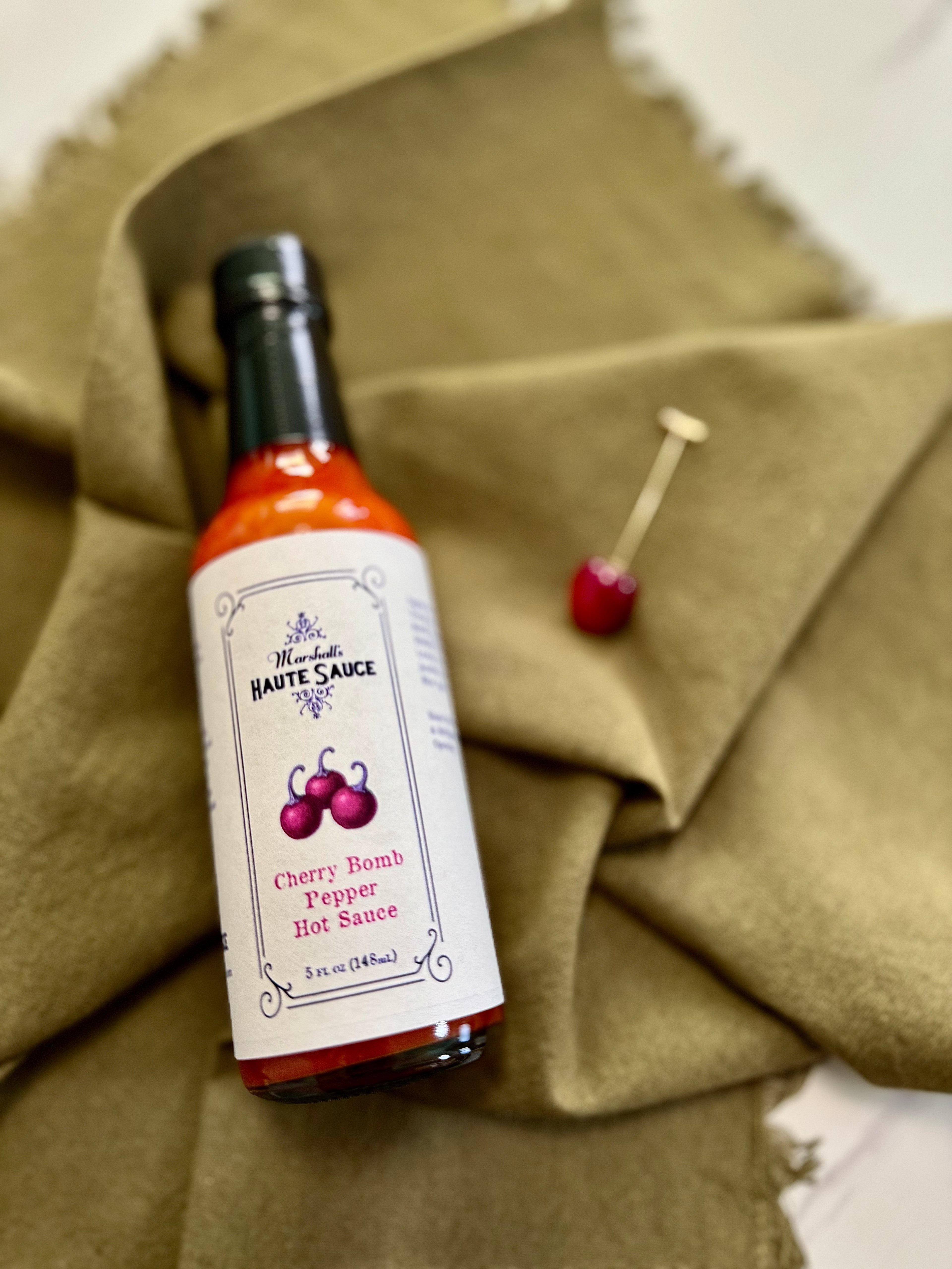 Cherry Bomb Pepper Hot Sauce bottle on olive green fabric, showcasing bold flavors and vibrant cherry design.