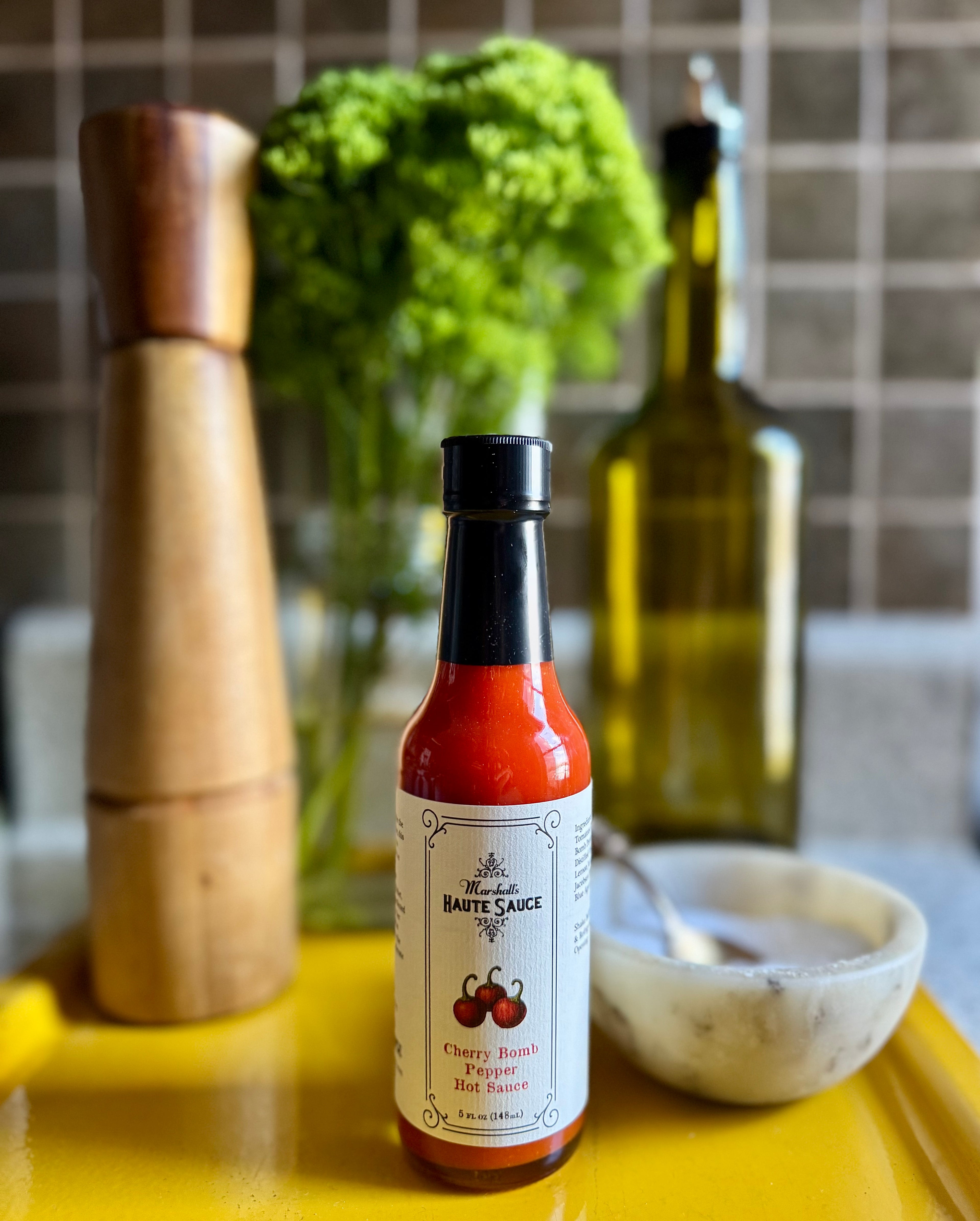 Cherry Bomb Pepper Hot Sauce bottle on a vibrant yellow tray with cooking ingredients in a kitchen setting.